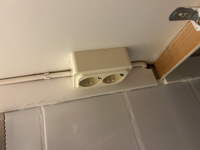 Gammalt 2-vägsuttag under köksskåp, med vit sladd som går igenom väggen, placerat ovanpå kakelplattor och intill en trälist.