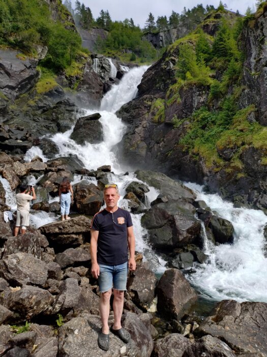 Man står framför ett vattenfall i en bergig skogsmiljö nära Geilo, Norge, med grönskande träd och stenar runt omkring.