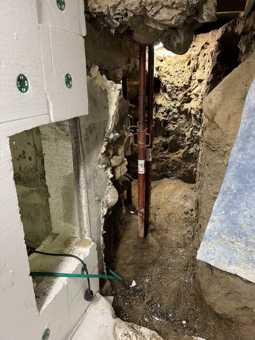 Källarutrymme med utsatt jord och murbruk, en stödjande metallstång samt vita isoleringsplattor på väggarna. En elkabel syns i hörnet.