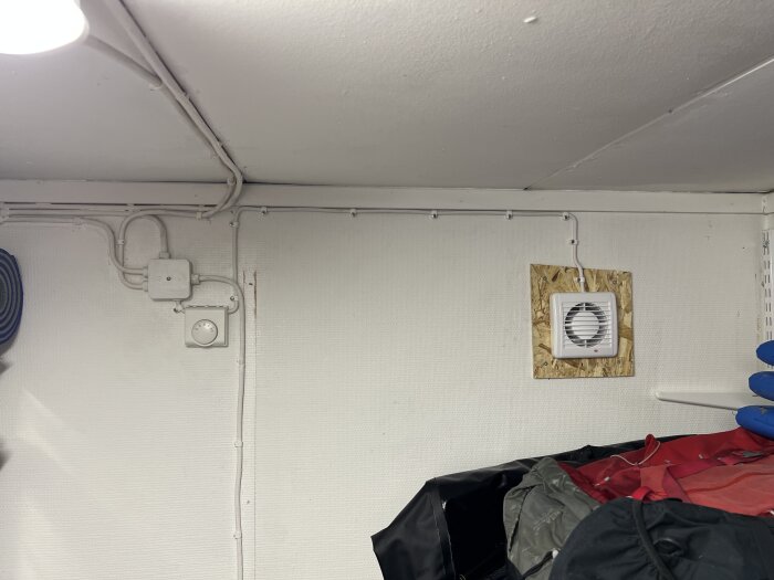 Badrumsfläkt med termostat installerad i förråd med två frysar, omgiven av vägg och förvaringsmaterial, för att sänka temperaturen.
