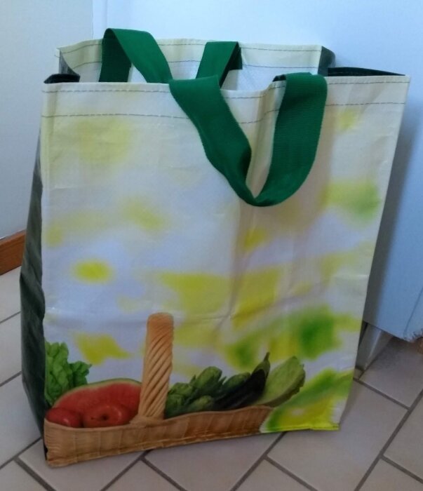 Kraftig Lidl-kasse i tjock plast med gröna handtag och bild på en korg med grönsaker tryckt på sidan. Står på ett kaklat golv.