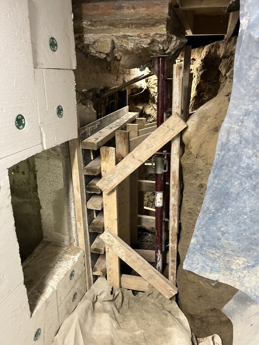 Källarrenovering under process med trästöd och stålbjälkar på plats, sammanfogande isoleringsmaterial, och påbörjad förstärkning av väggar.