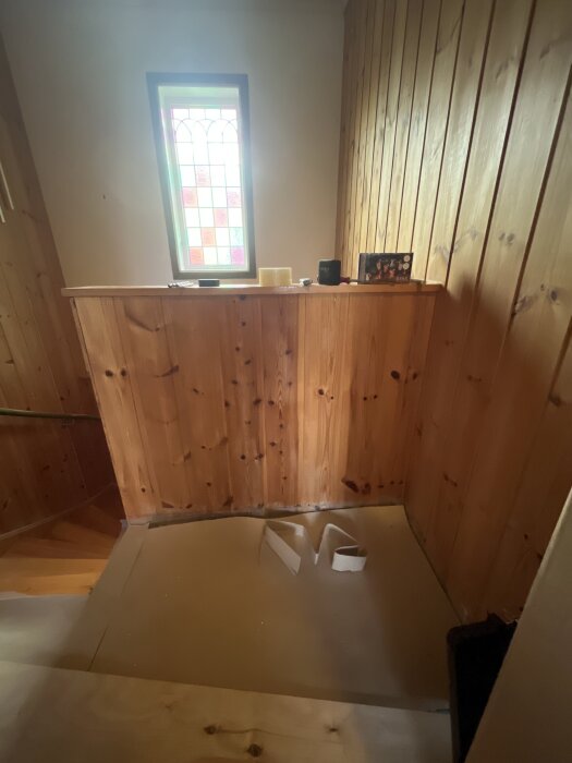 Träpanelvägg med ett avfasat fönster ovanför en trappa, förberett för att byta trappräcke.
