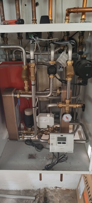 Värmesystemcentral med rörkopplingar, ventiler, pump och en elektronisk display monterad för övervakning.