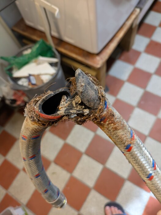 Sprucken anslutningsslang till varmvattenberedare, med trasig yttre väv och synlig insida, ligger på rutigt golv i en källare.