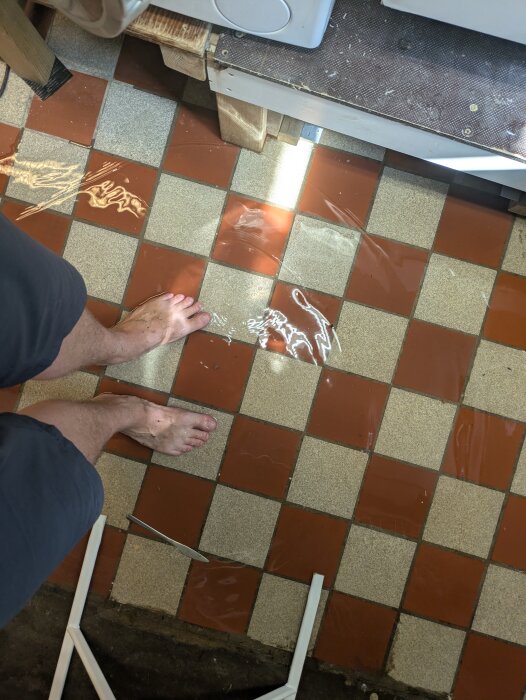 Fötter i vattenpöl på rödmönstrat källargolv. Vattenansamling efter sprucken varmvattenberedarslang.