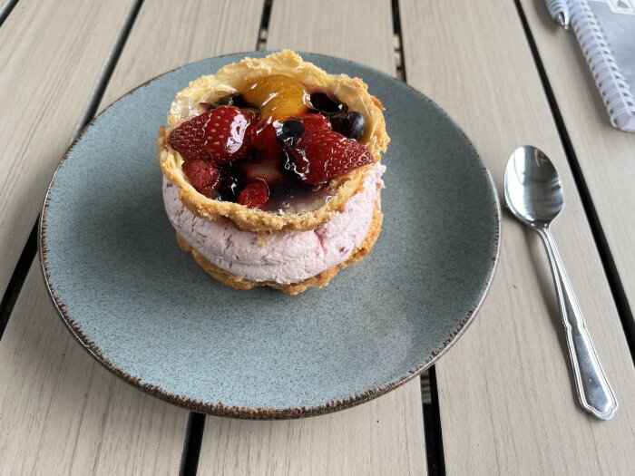 Bakelse med blåbärsmousse toppad med jordgubbar och andra bär på en blå tallrik, bredvid en sked och en anteckningsbok på ett bord.