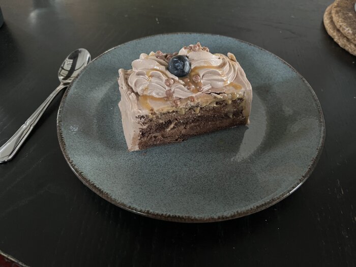 Bakelse med choklad och blåbärsmousse på en tallrik, dekorerad med blåbär och vispad grädde, bredvid en sked på brunt bord.