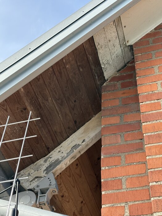 Fasaddetalj med synliga takstolar och råspont på ett äldre hus, viss färg är flagnad och råsponten verkar delvis omålad, intill rött tegel.