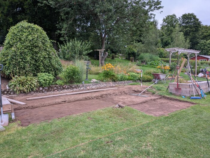 Grävt utomhusområde med trädgårdsredskap, redo för altanbygge; omgiven av gröna buskar och gräs.