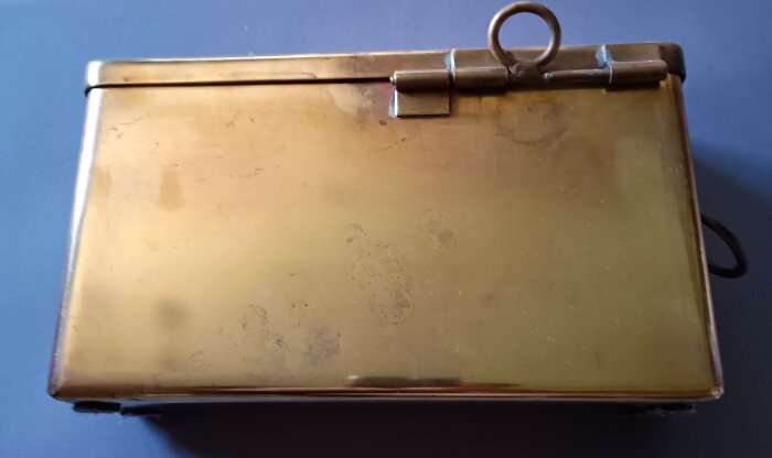 Ammunitionslåda i mässing med stängd lock och ring för öppning, fotograferad ovanifrån.