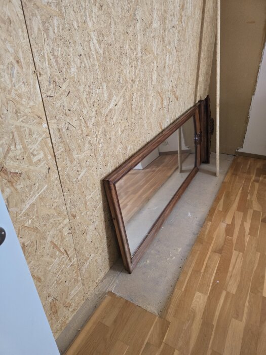 Masonitvägg som renoveras, isolering tillagt, spegel med träram står lutad mot väggen på trägolv.