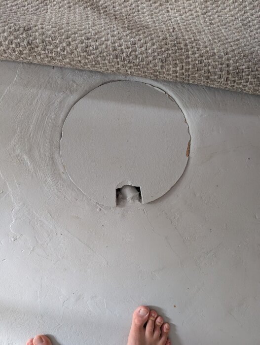 Gammal golvbrunn i källarrum med fläckad betongyta och upprullad matta intill. Fötter syns i bildens nederkant.