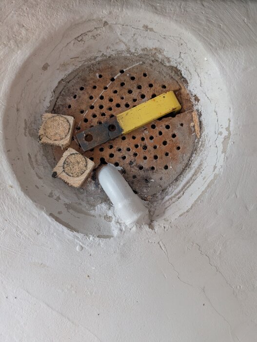 Gammal golvbrunn i källare med rostig plåt och ett gult verktyg, omgiven av slät betong.