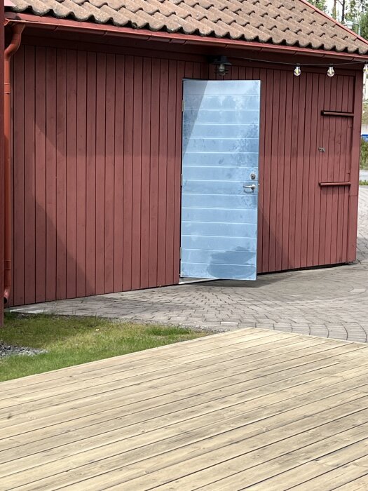 Rött garage med en blå dörr och tegelpannor på taket, omlämnat av stensatt gång och trätrall i förgrunden.