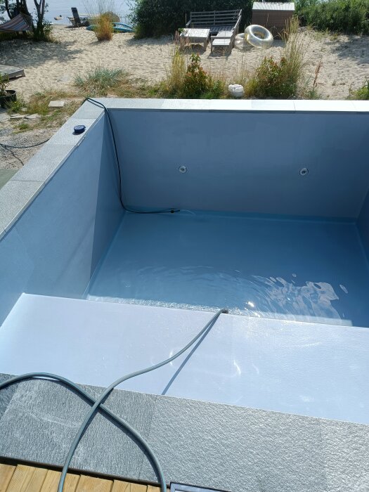 Fyllning av ett nästan tomt bassäng utan kantsten, med silvergrå färg som ser ljusblå ut. Vatten når 20 cm. Stranden syns i bakgrunden.