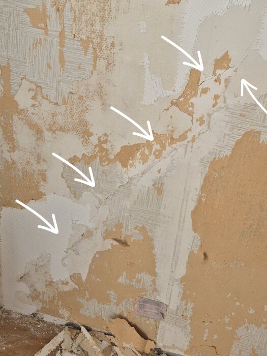Sprucken vägg med borttagen glasfiberväv och spackel, flera vita pilar pekar på sprickor under ytan.