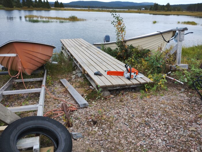 Brygga med ny trall, motorsåg och två båtar vid en sjö, omgiven av grus och växtlighet.
