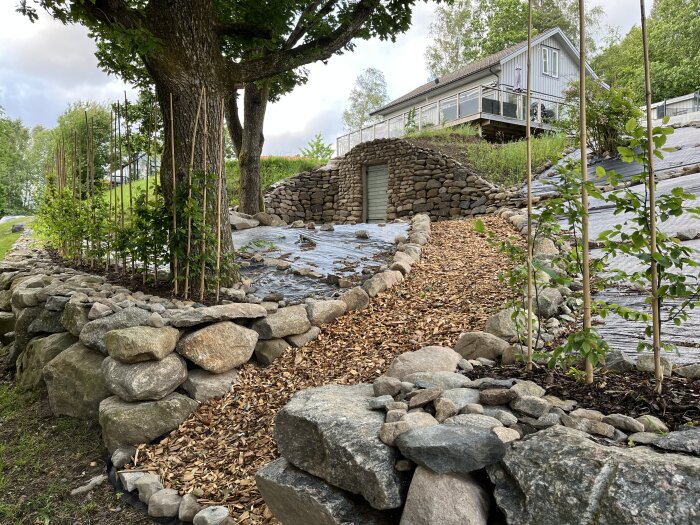 Stenmur omger en gångstig täckt med träflis, med ett trädhus i bakgrunden och växter längs kanterna.