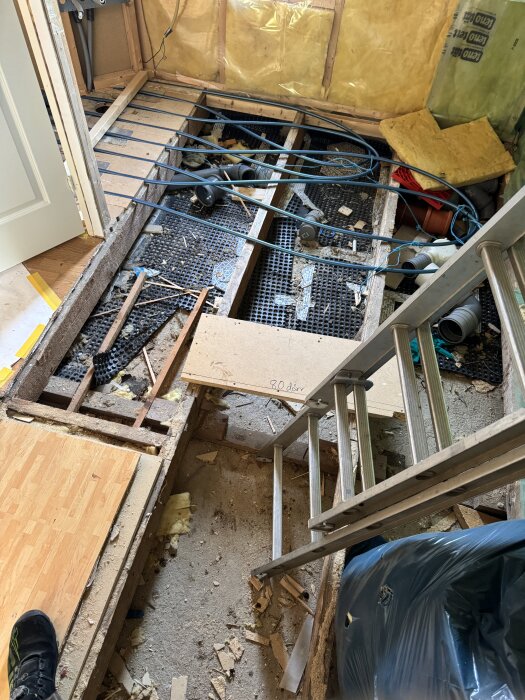 Badrumsrenovering med blottat golv, synliga golvvärmeslingor och isolering. En stege står lutad mot en vägg, och en regel markerar en planerad vägg.