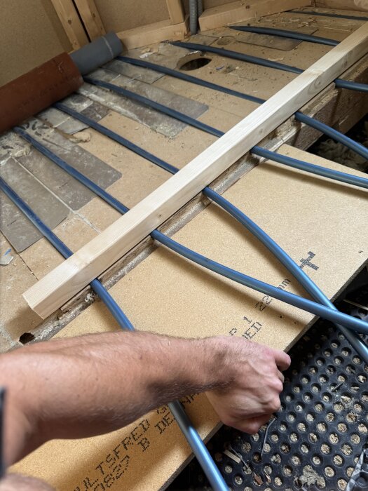En hand justerar golvvärmeslangar på ett öppet golv med en regel och spånplattor synliga i ett pågående badrumsbygge.