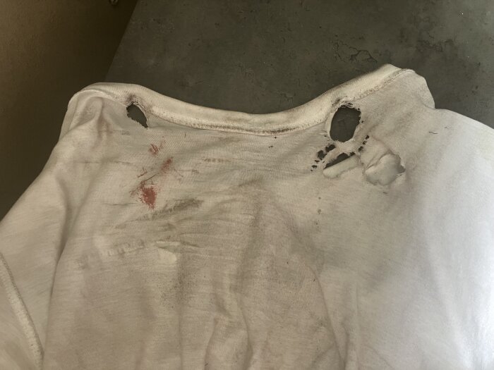 Vit t-shirt med flera hål och fläckar, troligen från ett verktygsrelaterat misstag.
