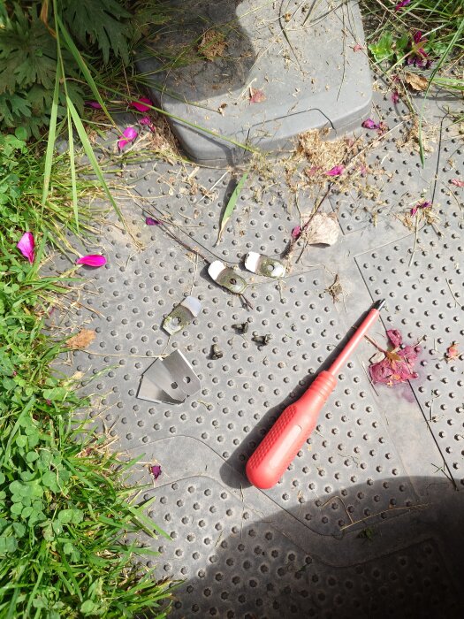 Gräsmatteknivar och en röd skruvmejsel på en gummimatta omgiven av gräs och blommor.