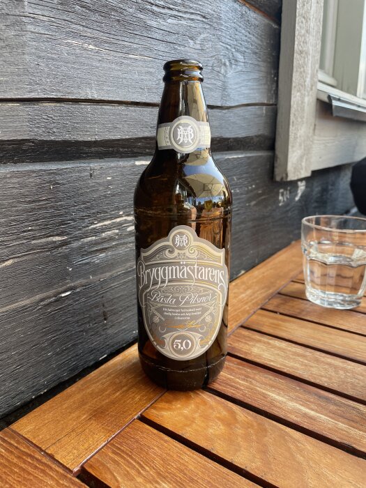 Flaska Bryggmästarens Bästa Pilsner placerad på ett träbord vid ett fönster med ett glas vatten bredvid.