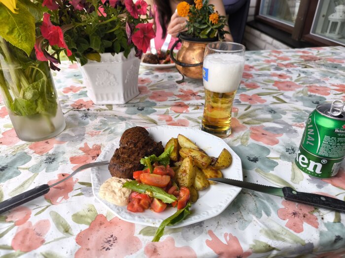 Grillad måltid med rostade potatisar, tomatsallad och kött på blommigt dukat bord, serverat med öl och läsk, blommor i bakgrunden.