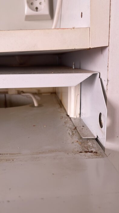 Del av köksskåp inuti, visar väggmonterad hylla med metallfäste och eluttag i bakgrunden. Hyllan verkar ha rengjorts.