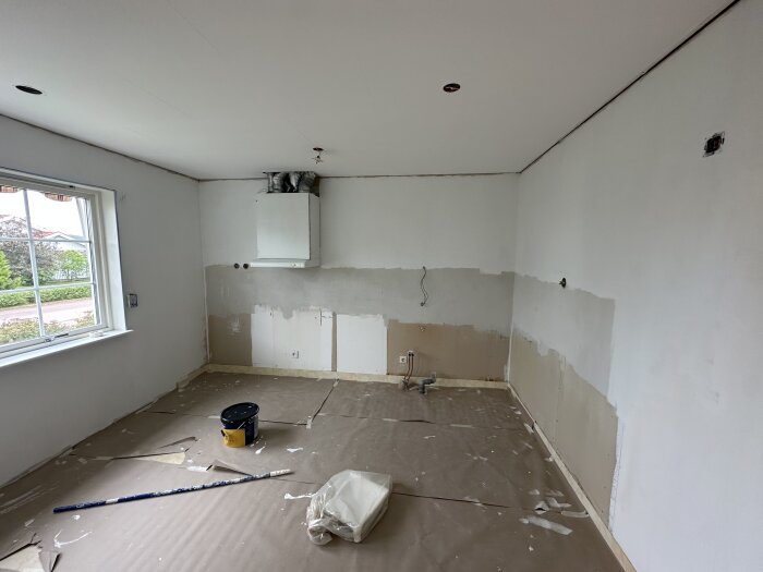 Renoverat kök med borttaget kakel, spacklade gipsväggar, fönster, värmepanna och golv täckt med skyddspapper. Pensel och färgburk synliga.