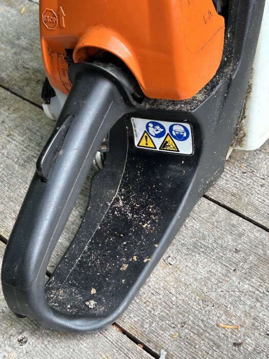 Närbild på en orange motorsåg med några smutsfläckar och varningssymboler på handtaget, placerad på ett träunderlag.