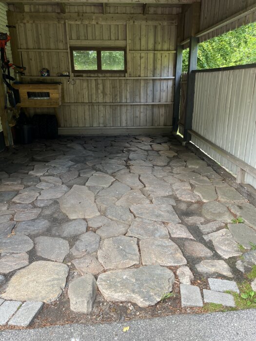 En carport med ojämnt stengolv, omgiven av träväggar och växtlighet.
