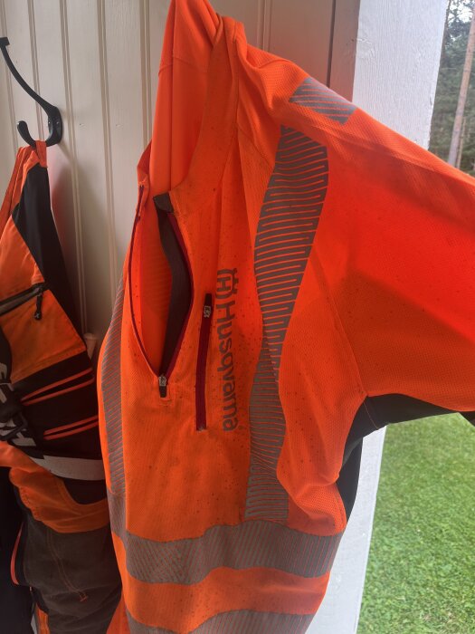 Orange Husqvarna huggarblus med reflekterande ränder hänger på en krok inomhus.