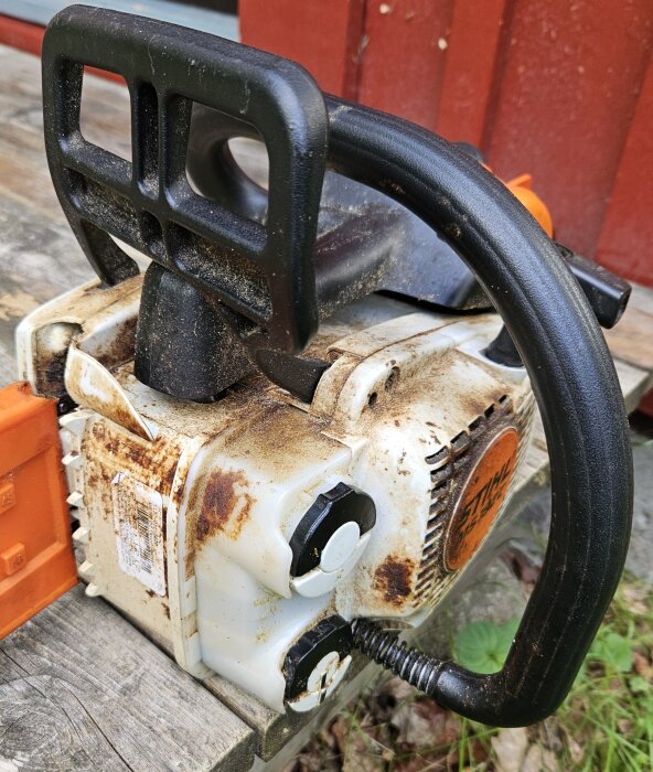 Sliten motorsåg med smuts och rost på avgassidan, ståendes på en träyta framför en röd vägg.