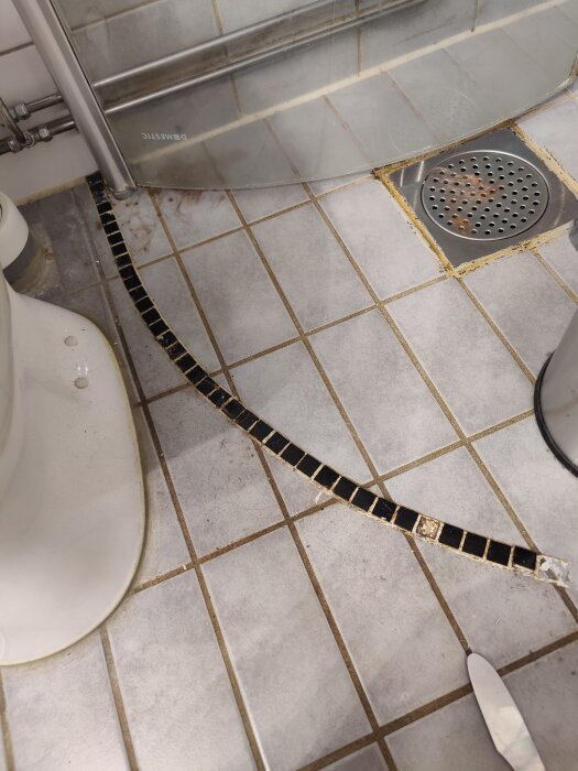 Svart mosaiklist i dusch med saknade bitar och dålig installation nära avlopp på badrumsgolv.