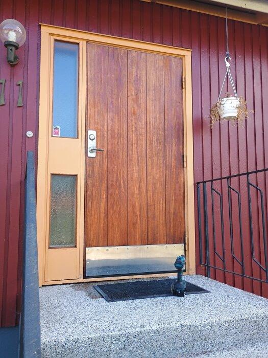 Träytterdörr med glasruta och borstat metallskydd, röd fasad, hängande kruka och en borrmaskin placerad på betongtrappa.