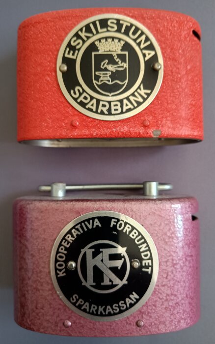 Röda och lila sparbössor med emblemen för Eskilstuna Sparbank och Kooperativa Förbundet Sparbanken, tillverkade av Låsbolaget i Eskilstuna.