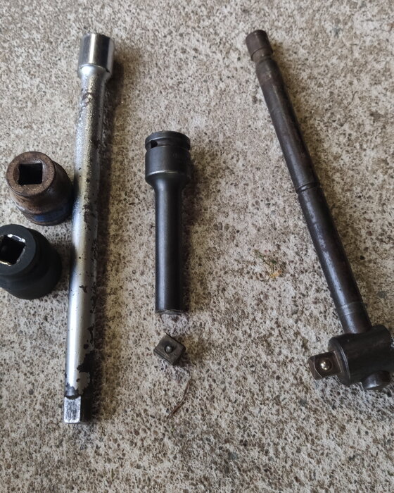 Olika storlekar av rostiga och slitna förlängare och hylsor ligger på ett betonggolv.