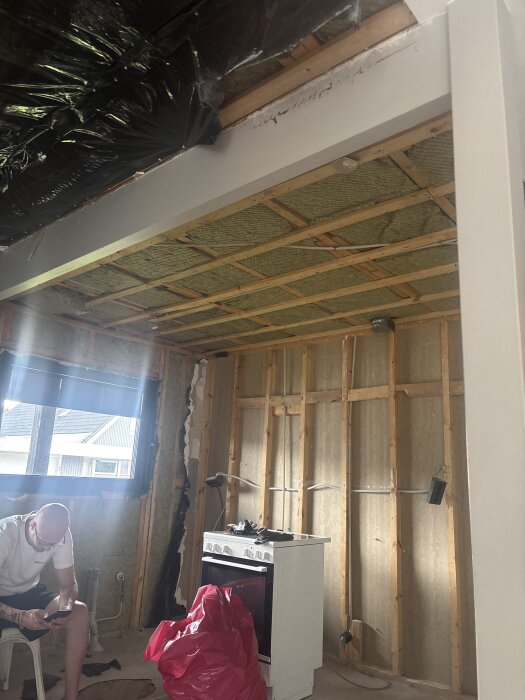 Ett rum under renovering med synliga reglar, takbalk, spis, man på stol, och rött föremål bredvid.