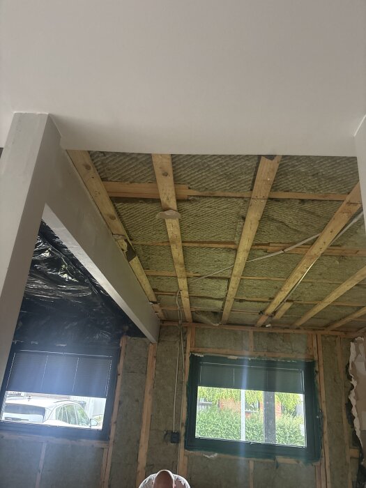 Tak med synliga träbjälkar och isolering i ett rum under renovering, med fönster och en synlig person nedanför.