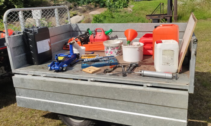 Olika verktyg och redskap ligger på en släpvagn, inklusive en domkraft, en motorsåg och flera dunkar, på en solig dag utomhus.