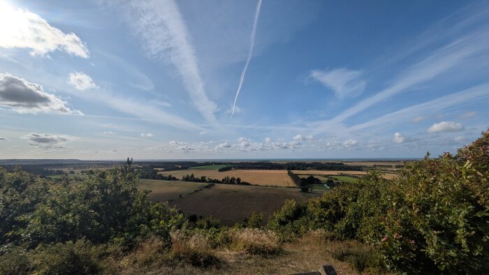 Utsikt över landskap med fält och skog under blå himmel med vita moln och kondensstrimmor.