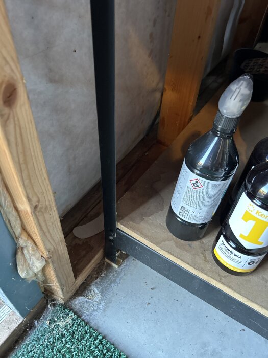 Insidan av en konstruktion med träreglar, en hylla med flaskor och isolering, samt en bit av ett grönt golv.