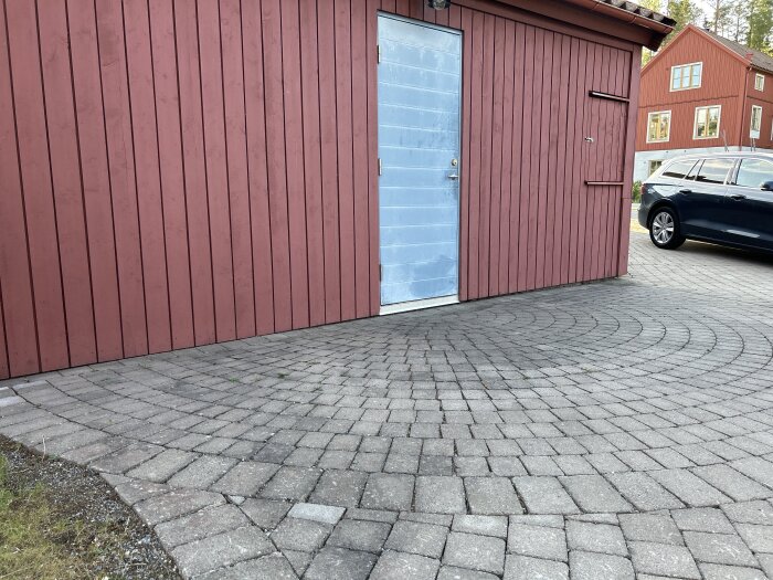 Röd trävägg med en blå dörr, stenbelagd gång framför och en parkerad bil till höger, samt ett rött hus i bakgrunden.