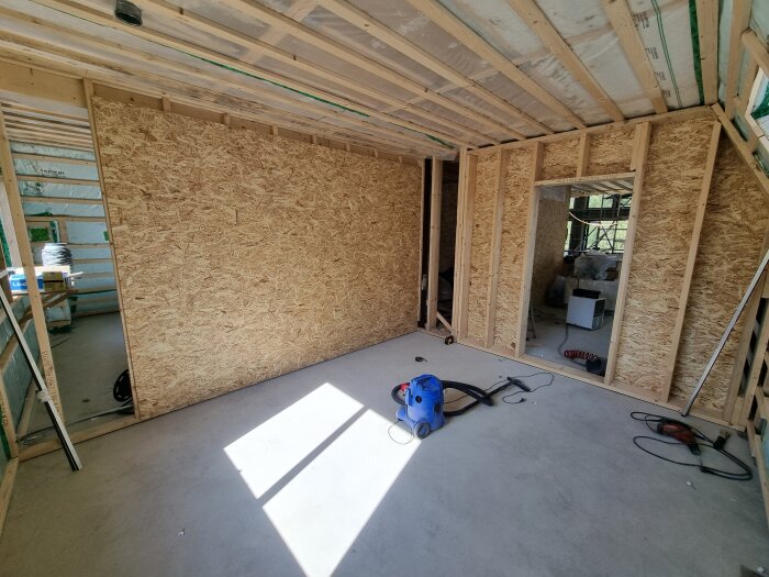 Rum under renovering med OSB-skivor och verktyg som dammsugare och borrmaskin på golvet, samt öppningar för framtida elinstallationer.