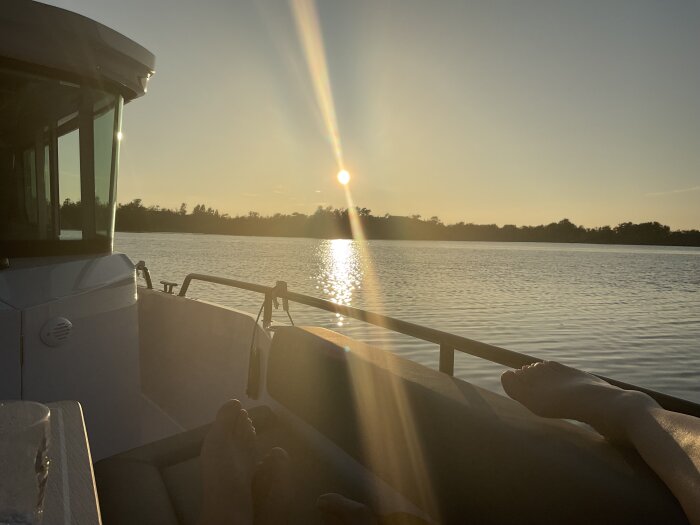Solnedgång över vatten sedd från en båt på St. Nassa, med fötter vilande på bänken och solstrålar som reflekteras i vattnet.