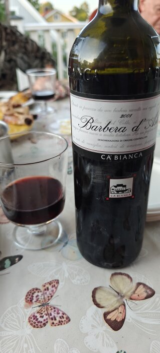 Flaska Barbera d'Asti vin med vinglas på bord med fjärilsmönster.