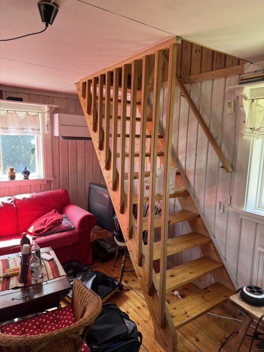 Trätrappa med lösa steg i ett vardagsrum med röda soffor och luftvärmepump.