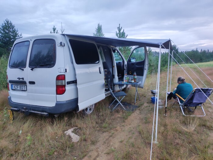 Vit skåpbil med uppfällt tak vid en lägerplats, bord och stolar utanför, mellan träd nära svenska gränsen, molnigt väder.